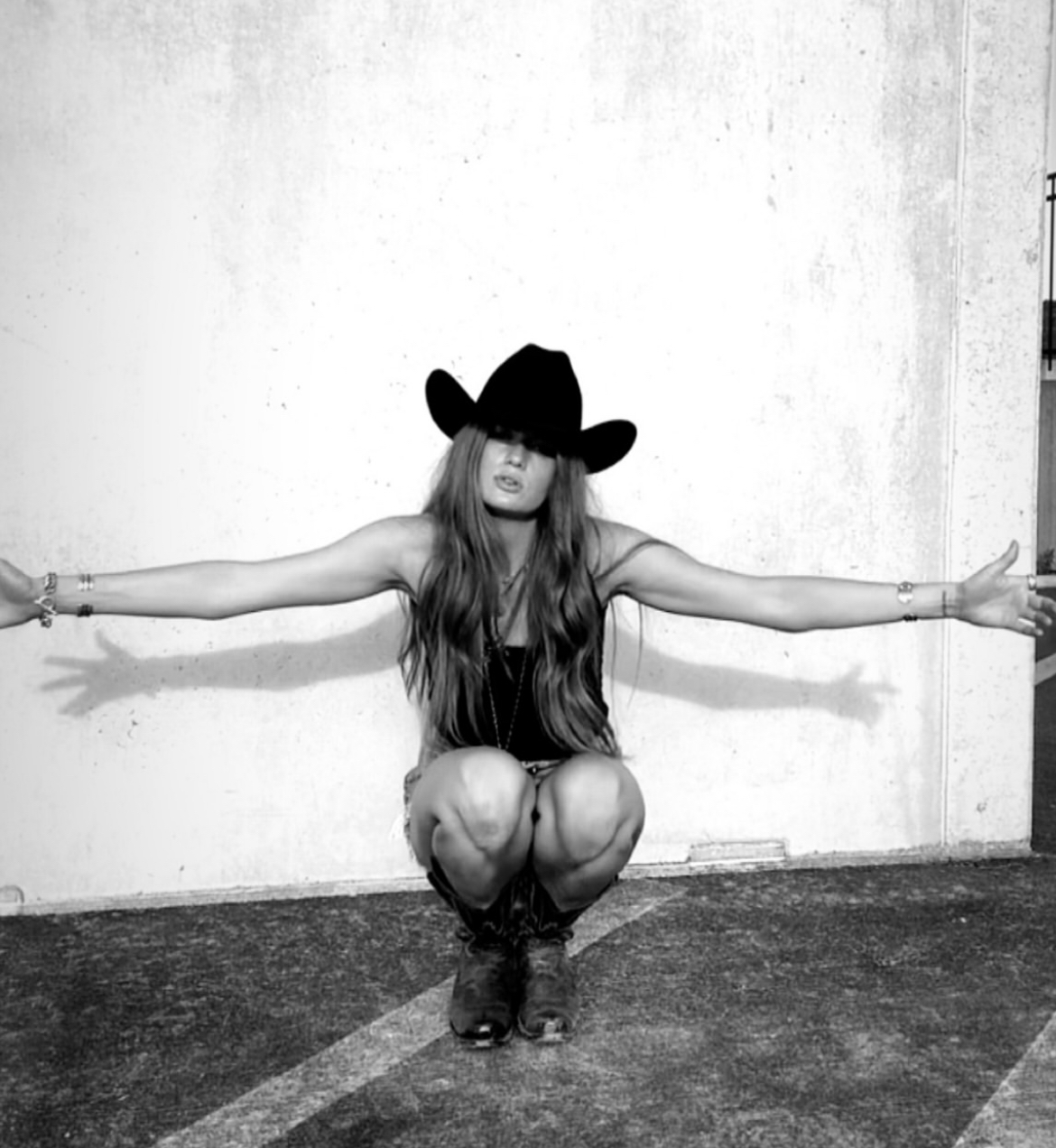 Woman crouching with arms outstretched, wearing a hat, long hair, and boots, against a plain wall background.