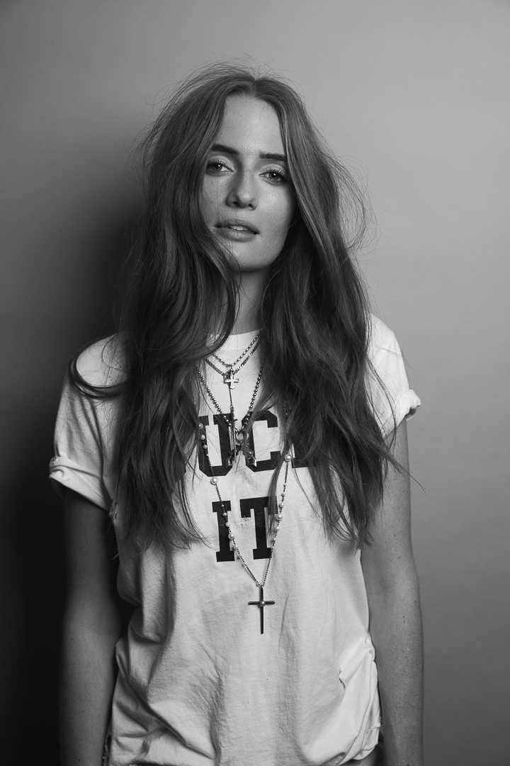 Young woman with long wavy hair wearing a T-shirt with text and necklaces, looking at the camera.
