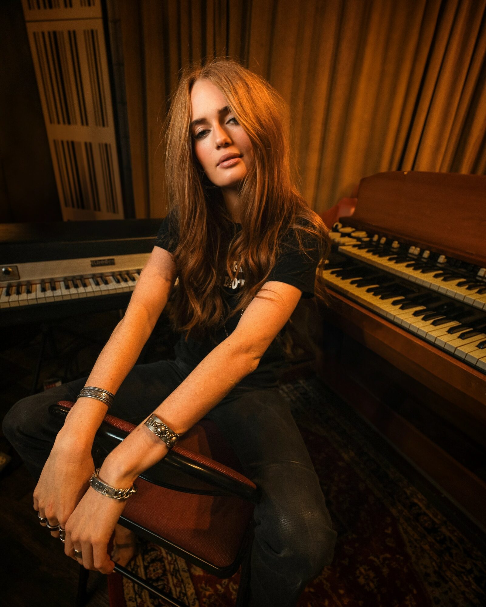 Young woman with long hair sitting on a bench in a music studio, surrounded by pianos and musical equipment.