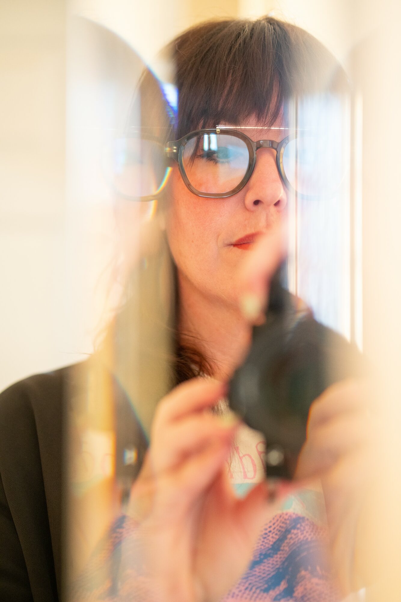 Person with glasses taking a photo through a mirror, reflection visible, holding a camera, indoors.