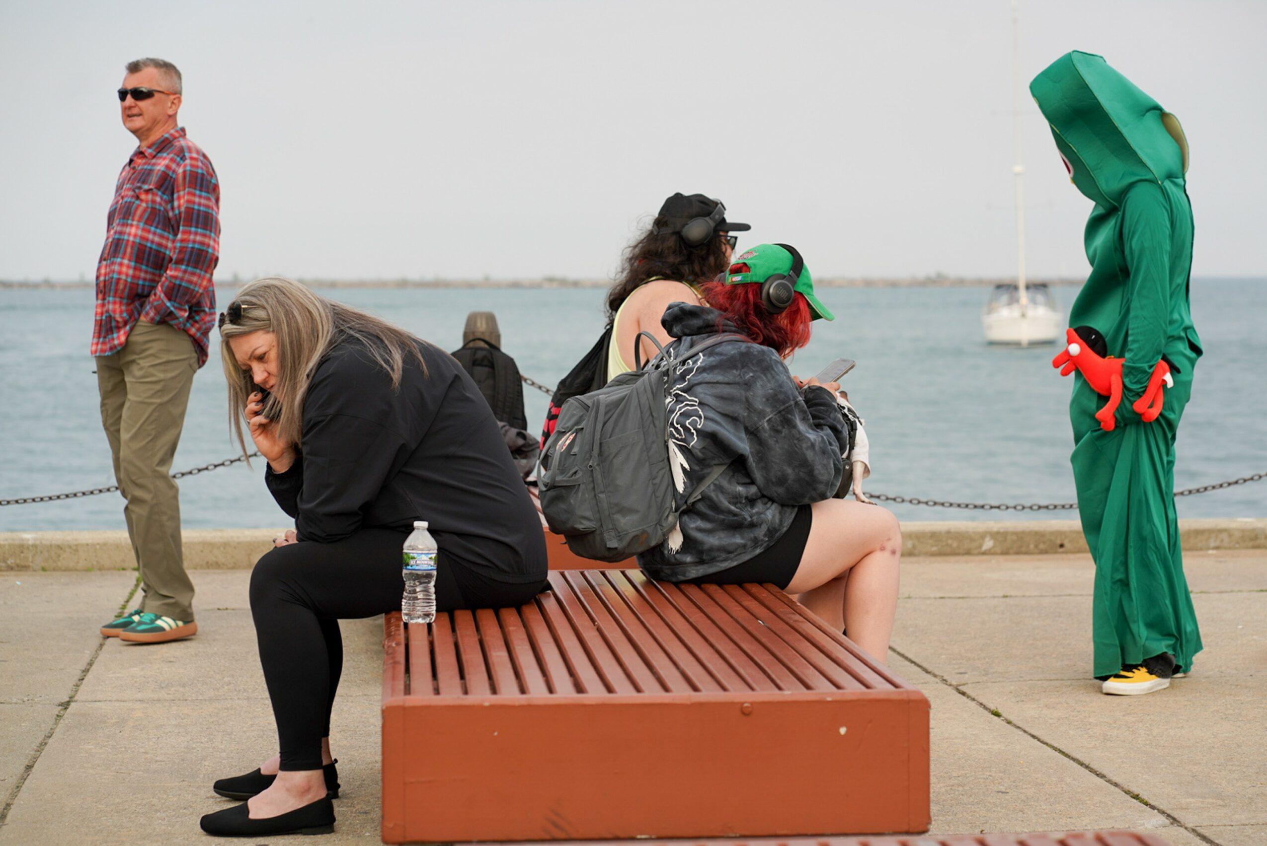 People sitting on a bench near water, one person in a green costume, others engaged in conversation, background with boats.