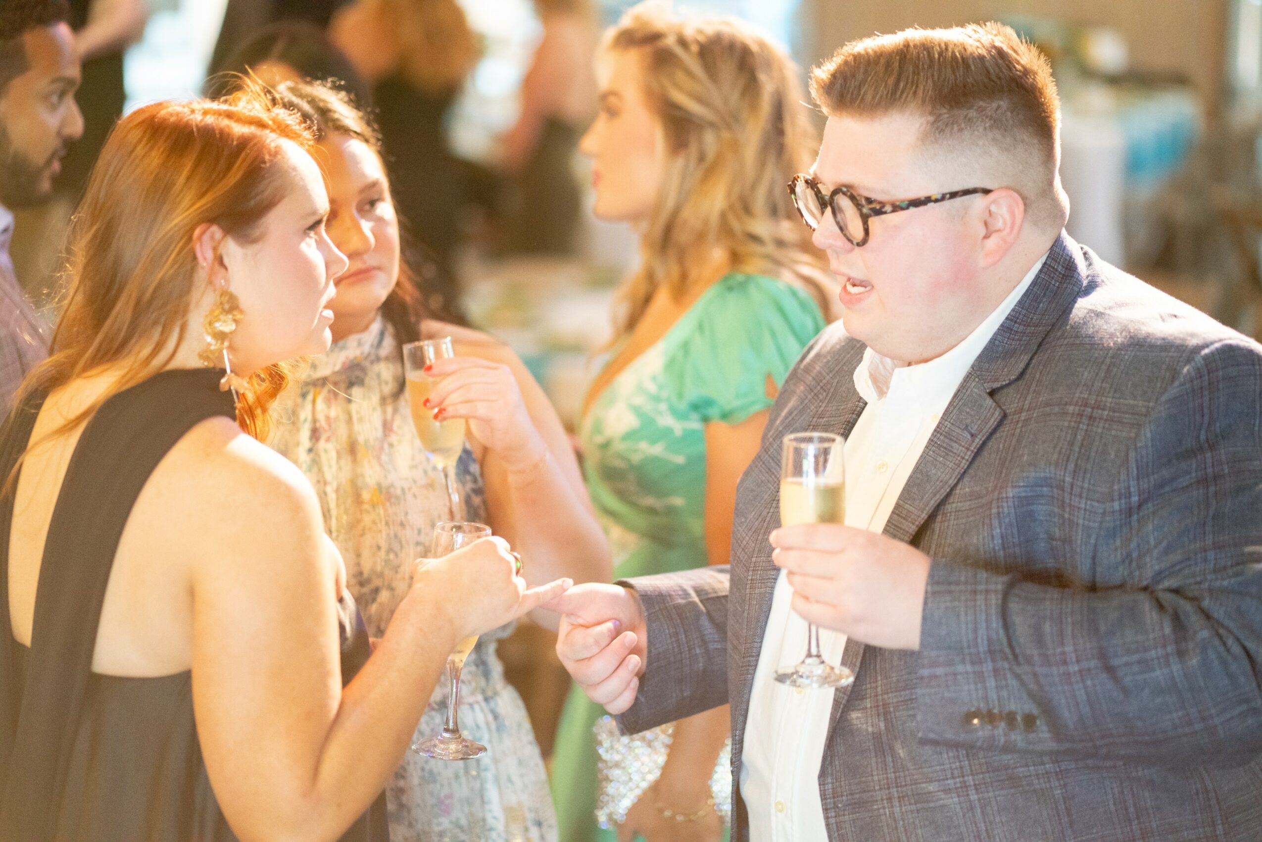 People at a social gathering, including a woman in a black dress and a man in a suit with glasses, shaking hands.