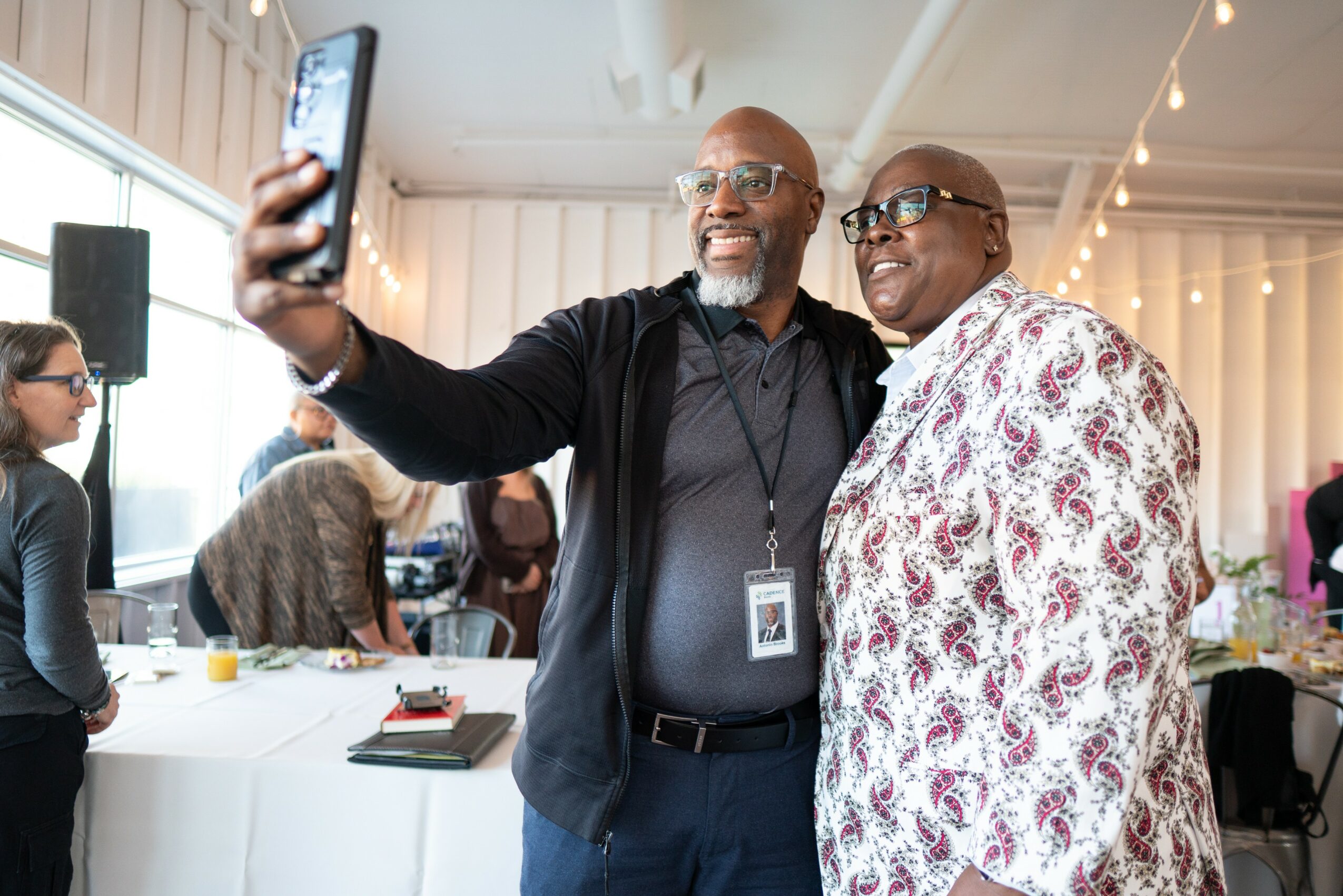Two men taking a selfie at a social event, smiling, with other people and tables in the background.