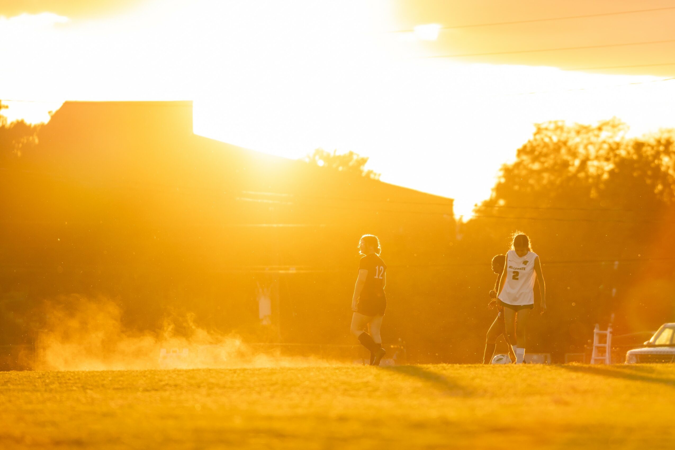 Two children walking outdoors during sunset with trees and buildings in background.