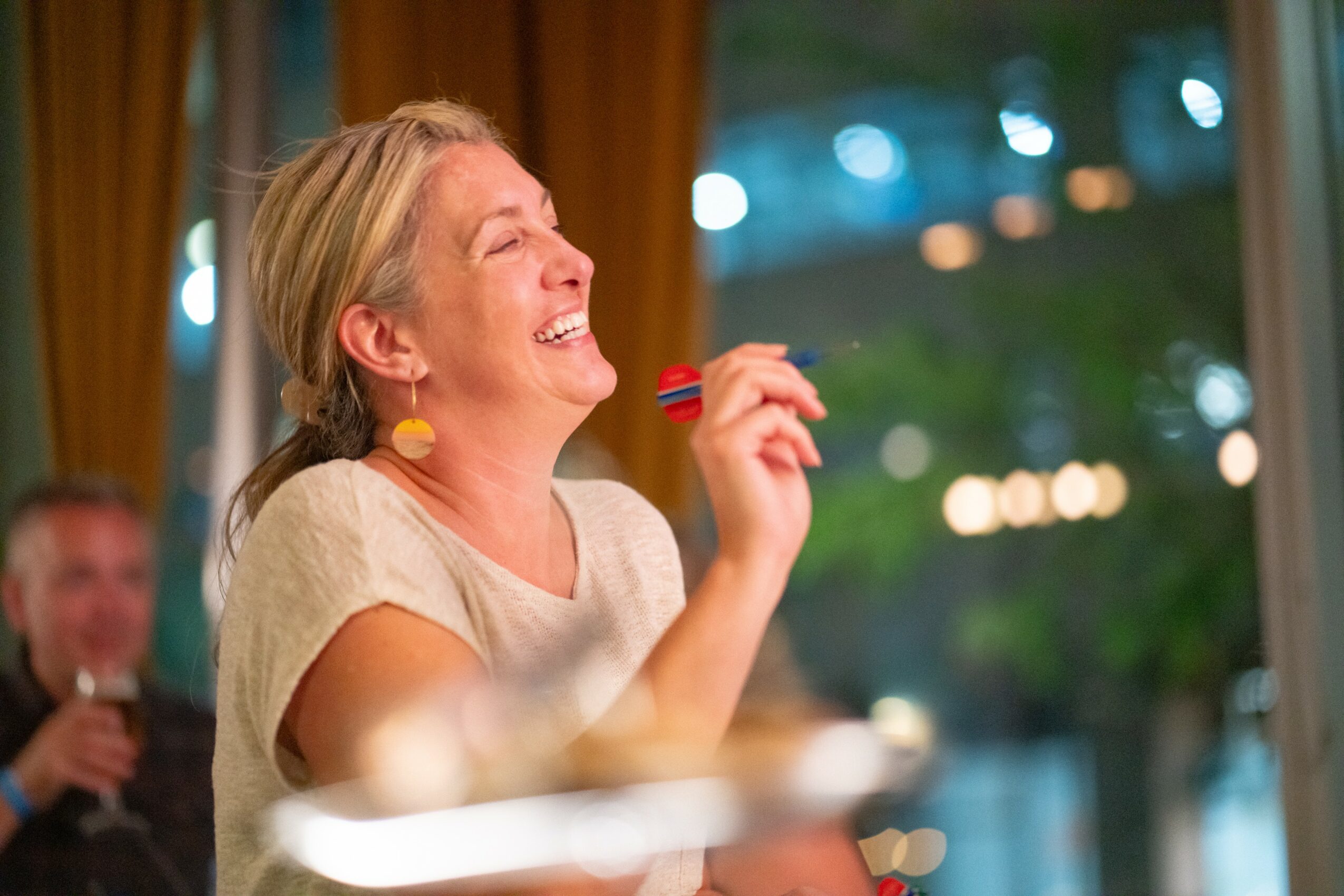 Woman laughing and holding a small object in a room with large windows and blurred outdoor lights.
