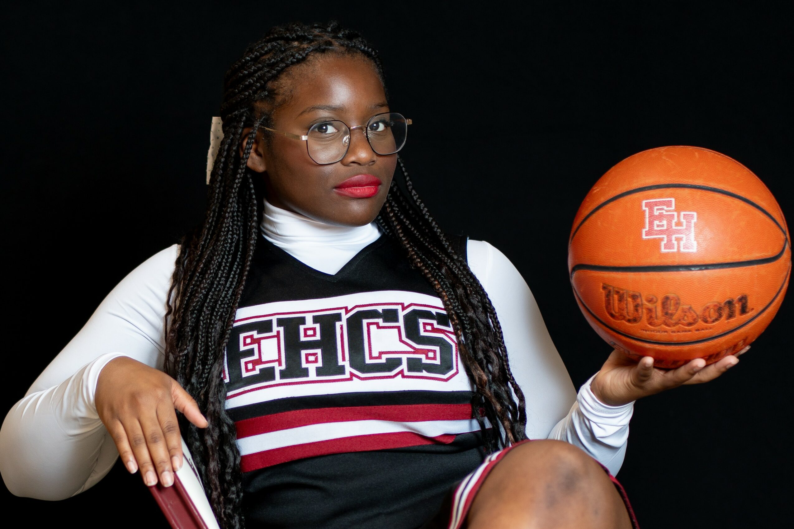 Woman in cheerleading uniform holding basketball against black background.