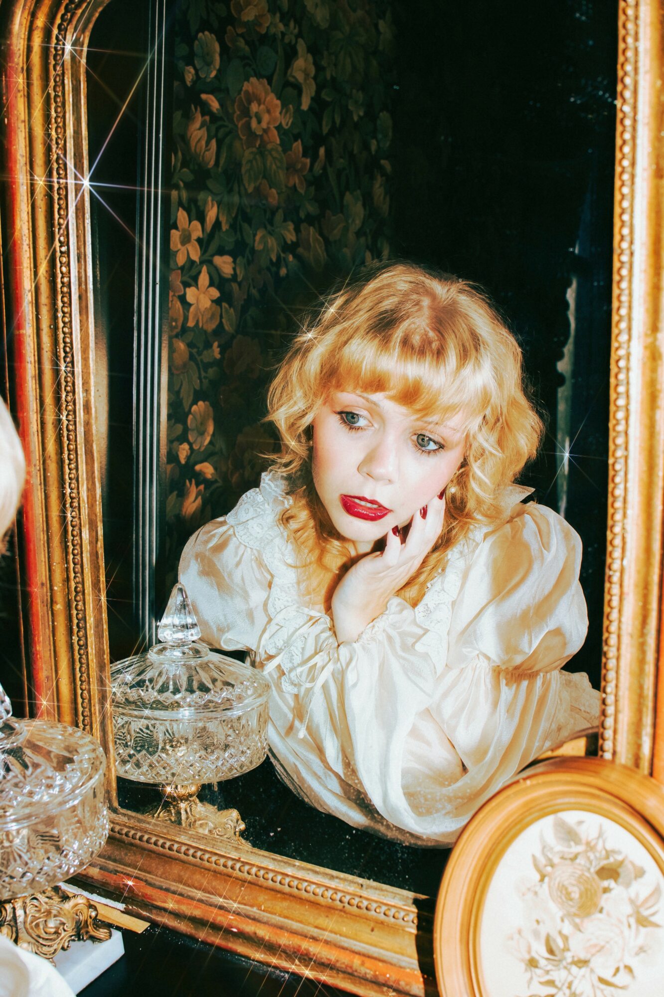 Young woman with curly blonde hair looking into mirror, resting her chin on hand, wearing a light-colored blouse.