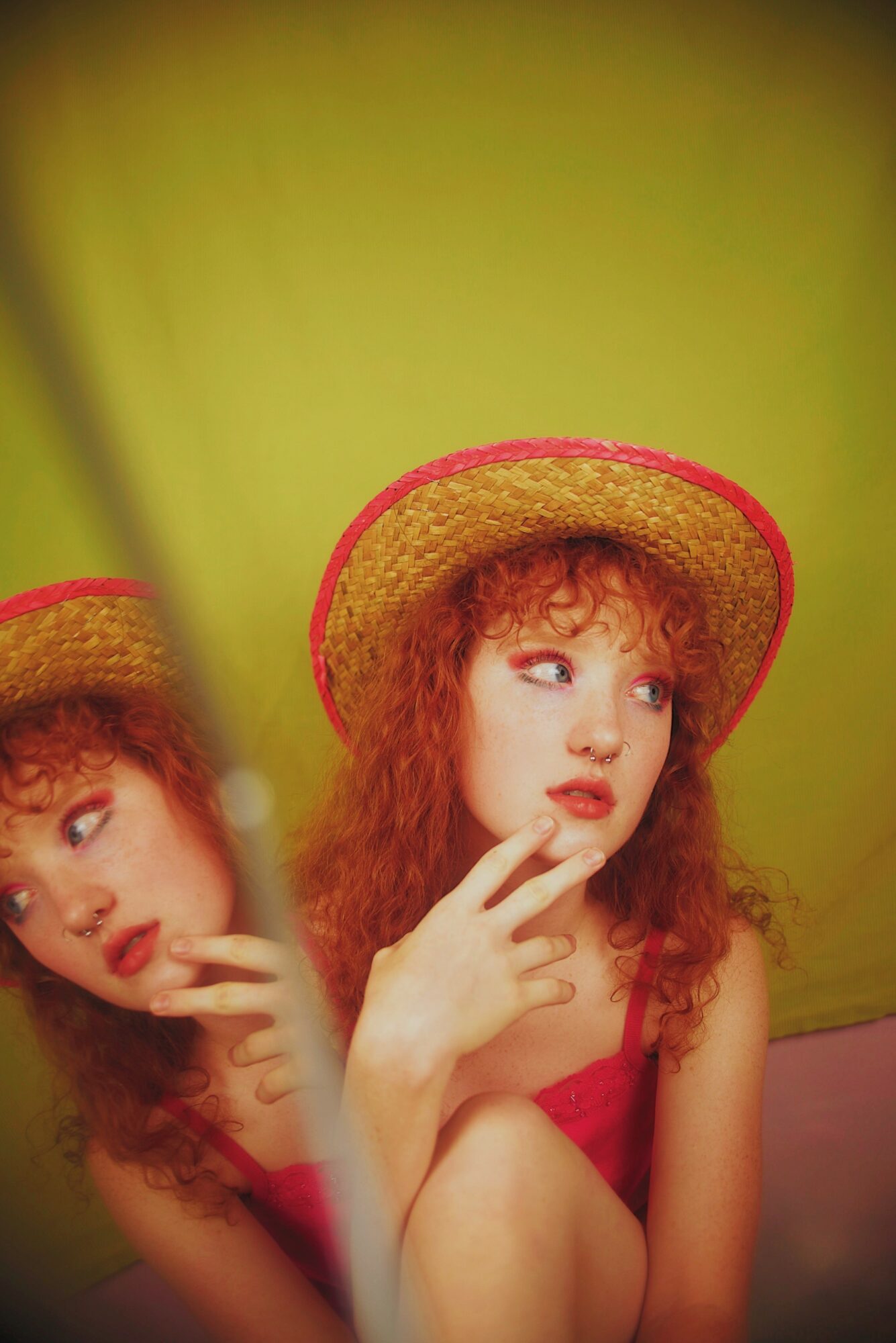 Young woman with curly red hair wearing a straw hat and pink top, touching her face, against a green background.