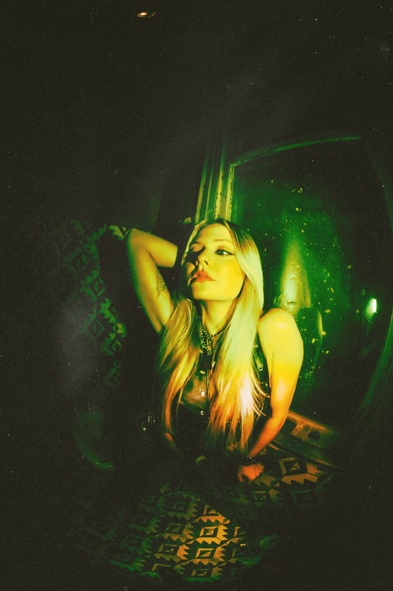 Young woman with long hair poses with one arm behind her head in a dark, green-lit environment.