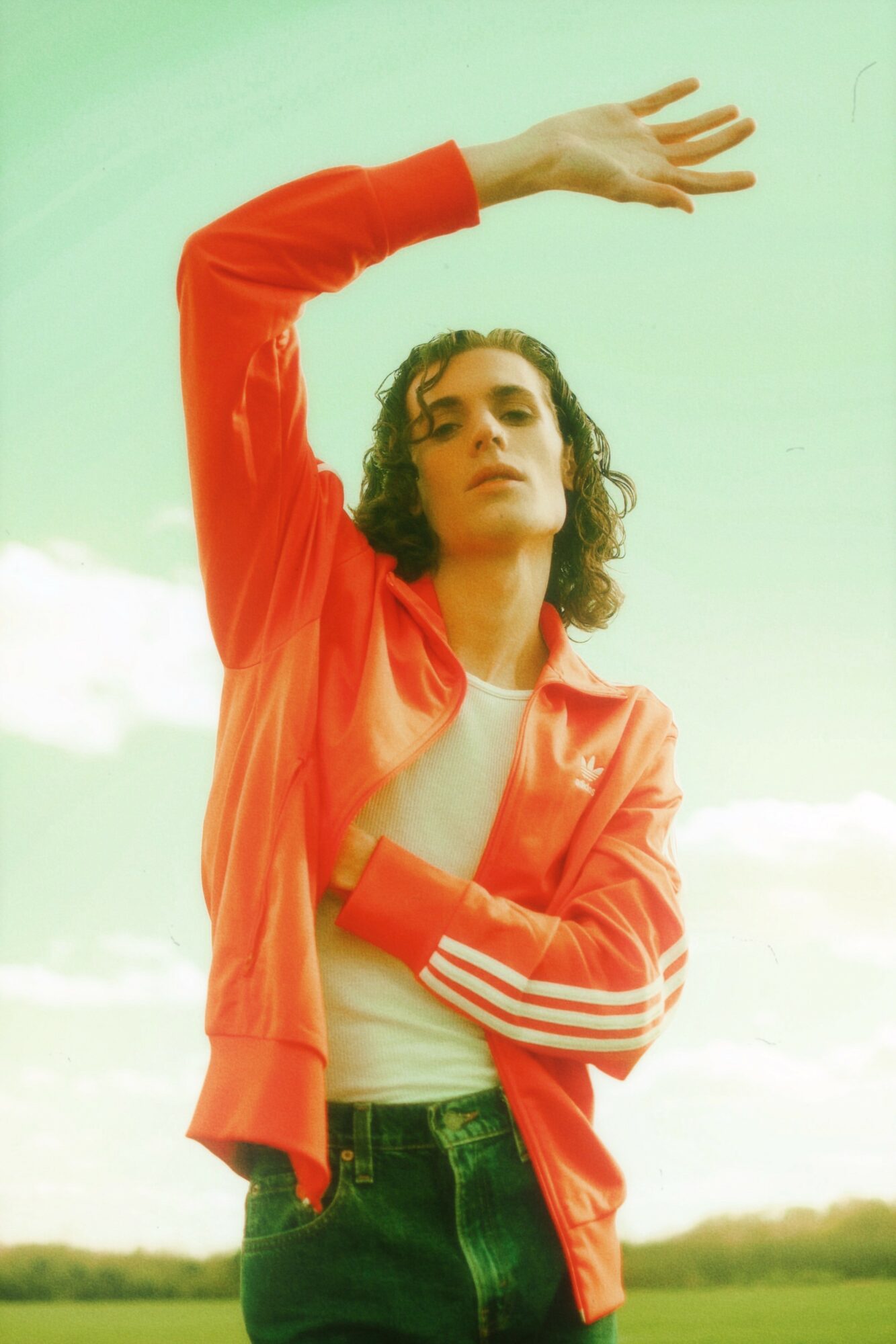 Woman with curly hair wearing a red jacket and white shirt, raising her arm outdoors against a cloudy sky.