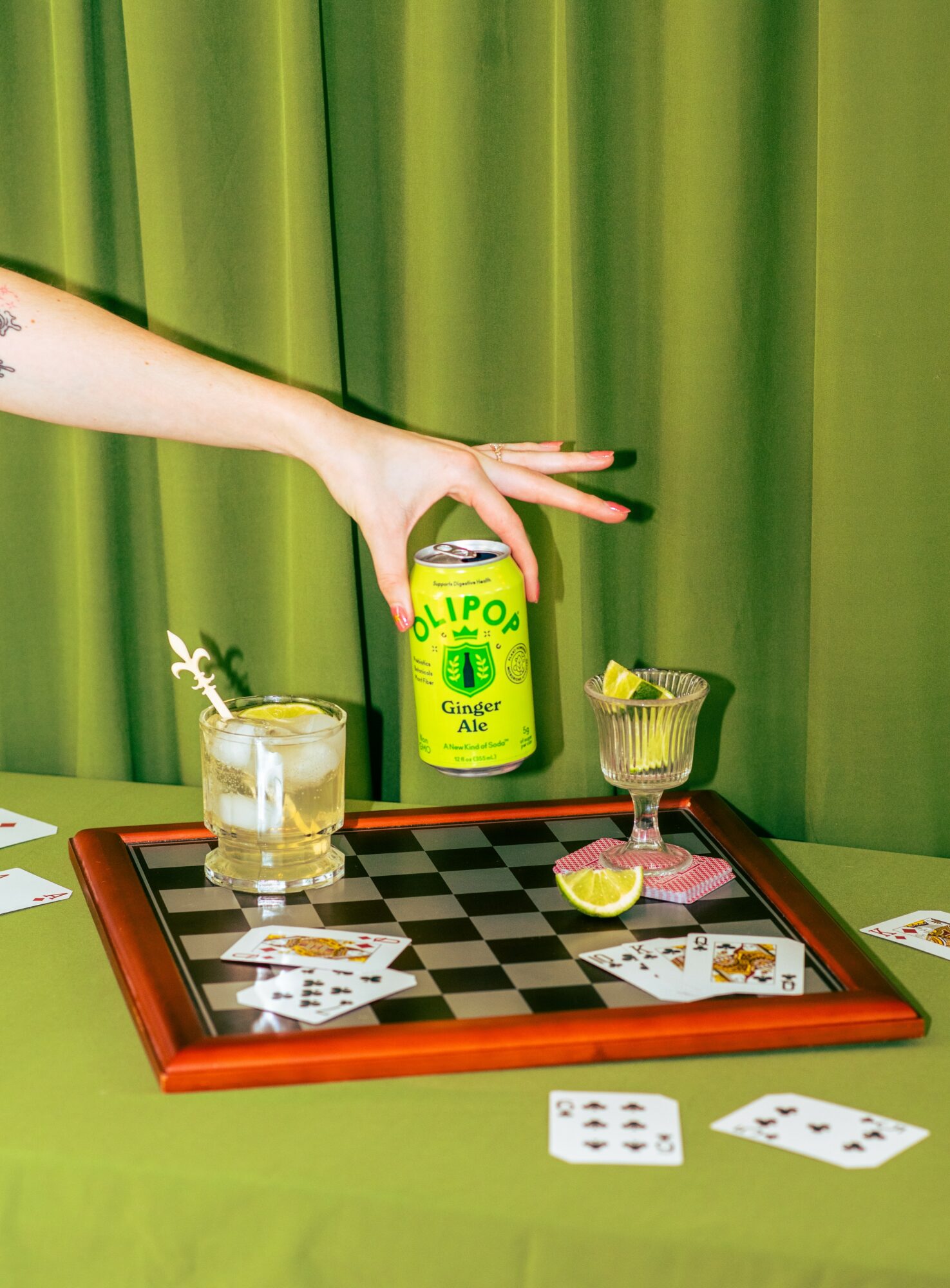 Hand reaching for a can of ginger ale on a checkered game board with drinks and playing cards.