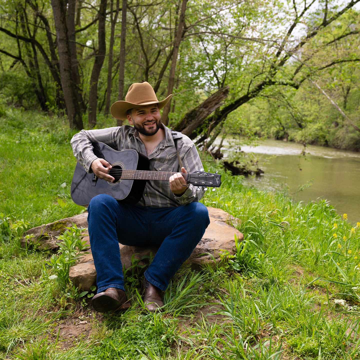 Man sitting on a rock playing guitar outdoors near a river, surrounded by trees and green grass.