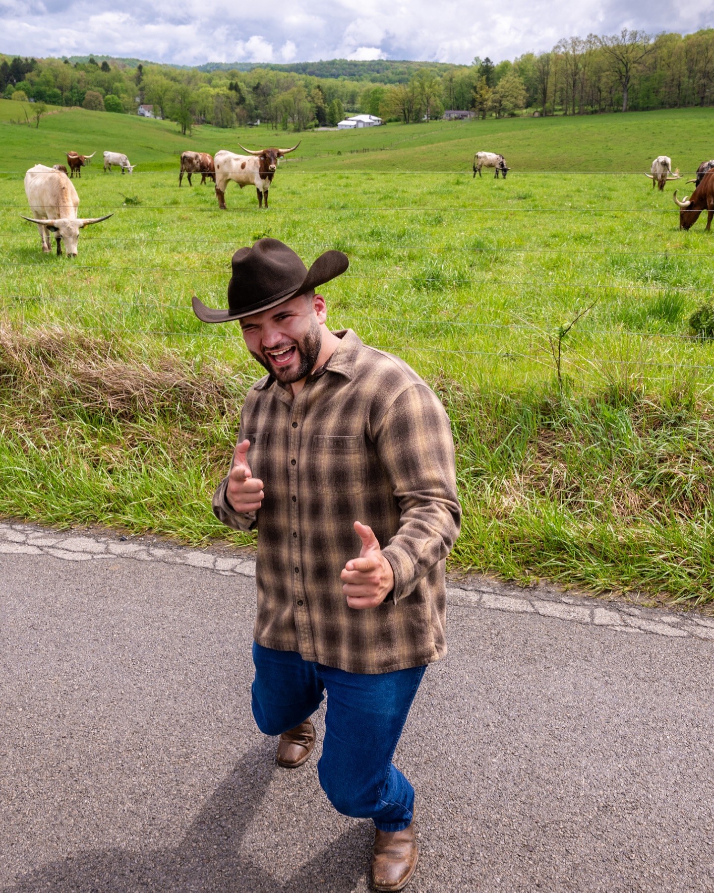 Person in plaid shirt and hat giving thumbs up on roadside with cows grazing in green field behind.