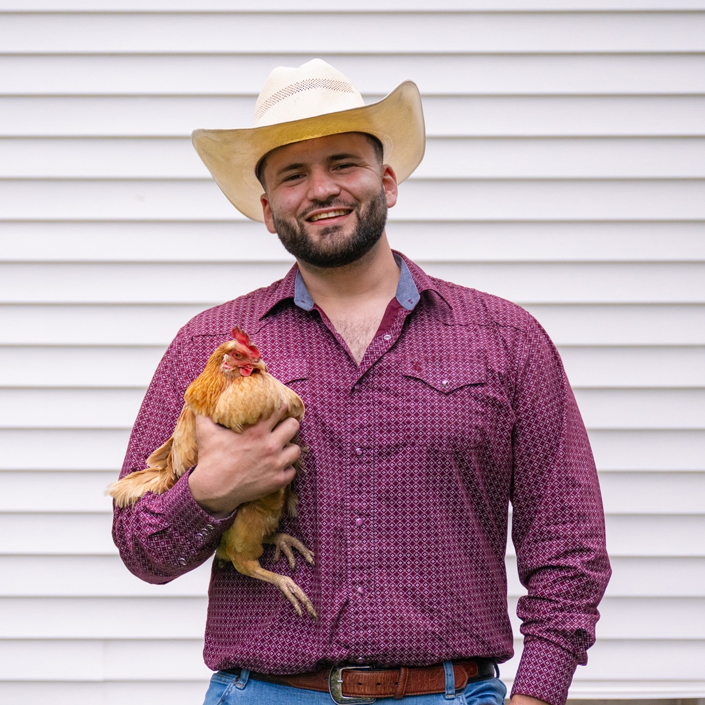 Man wearing cowboy hat holding a chicken, standing in front of a white horizontal siding wall.