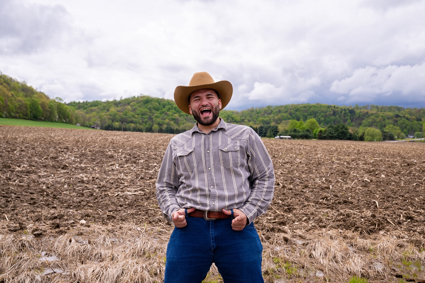 Man wearing a cowboy hat and striped shirt standing in a field with trees and cloudy sky in background.
