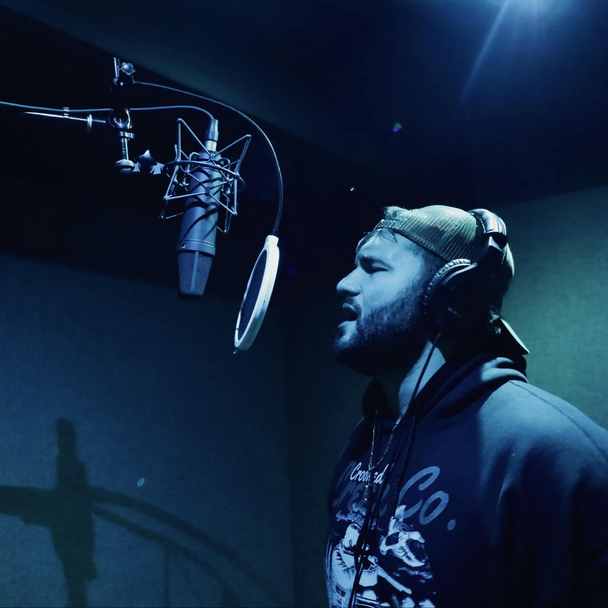 Man in headphones singing into a microphone in a recording studio with blue lighting.