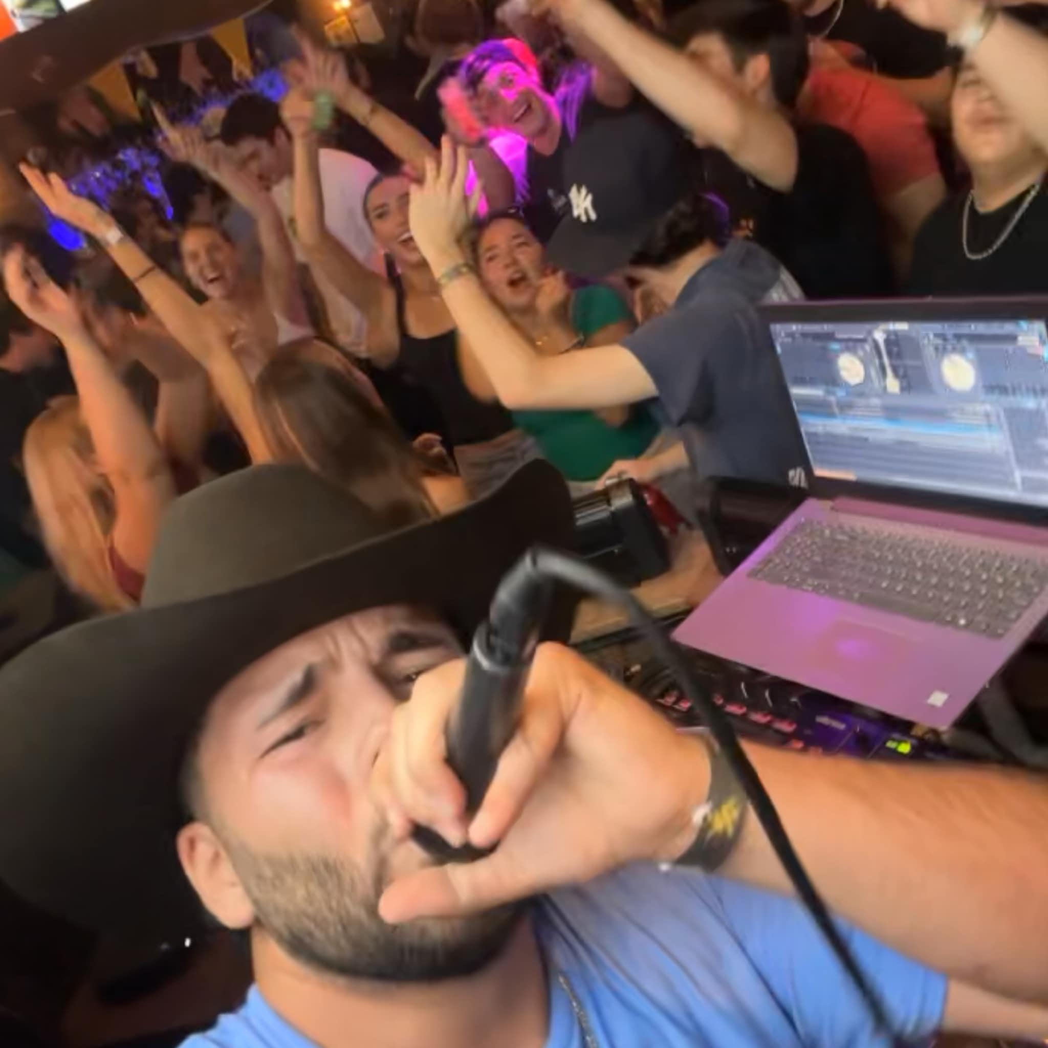 DJ with a hat singing into a microphone at a lively party with dancing crowd in background.