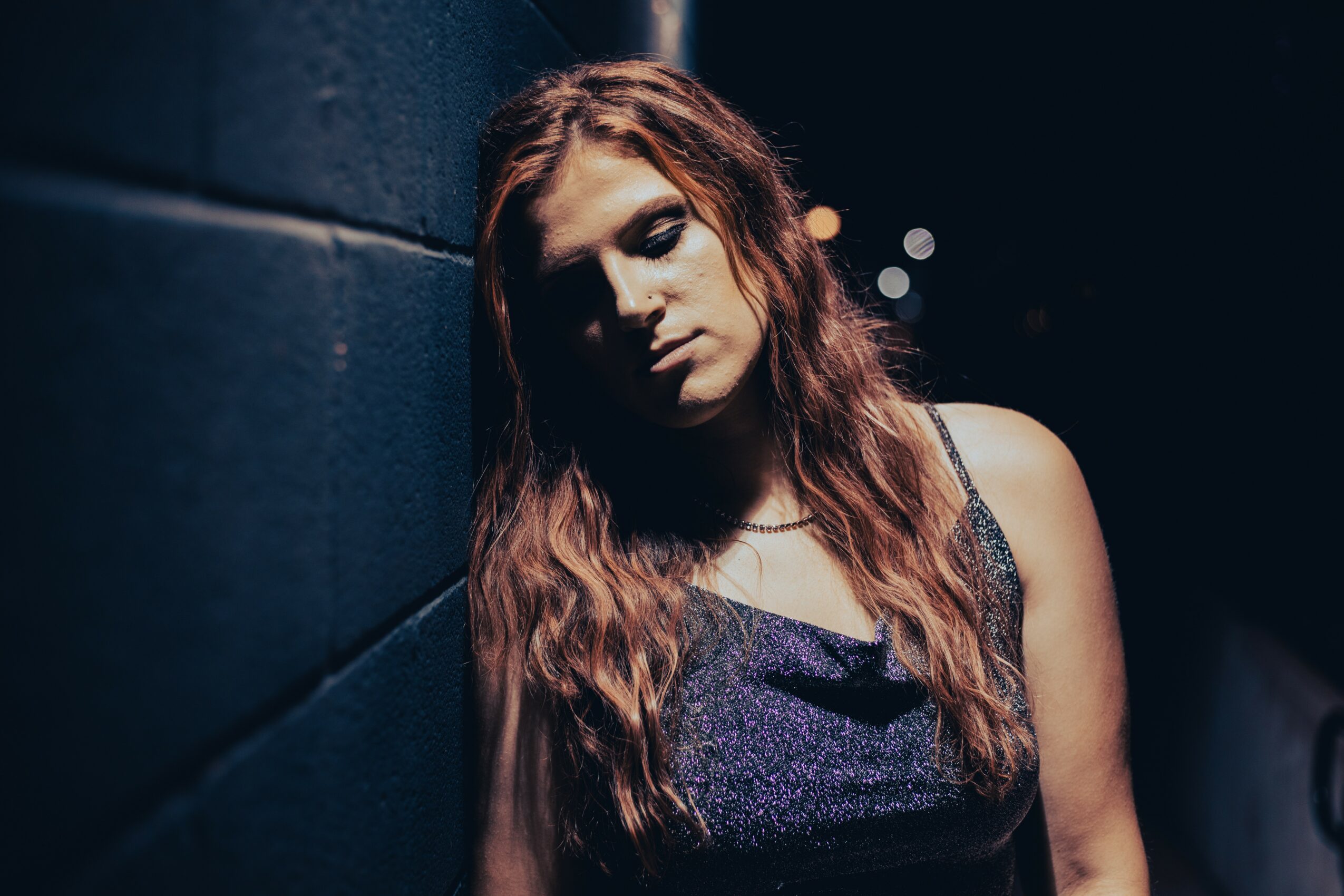 Young woman with long wavy hair leaning against a dark wall in a dimly lit environment.