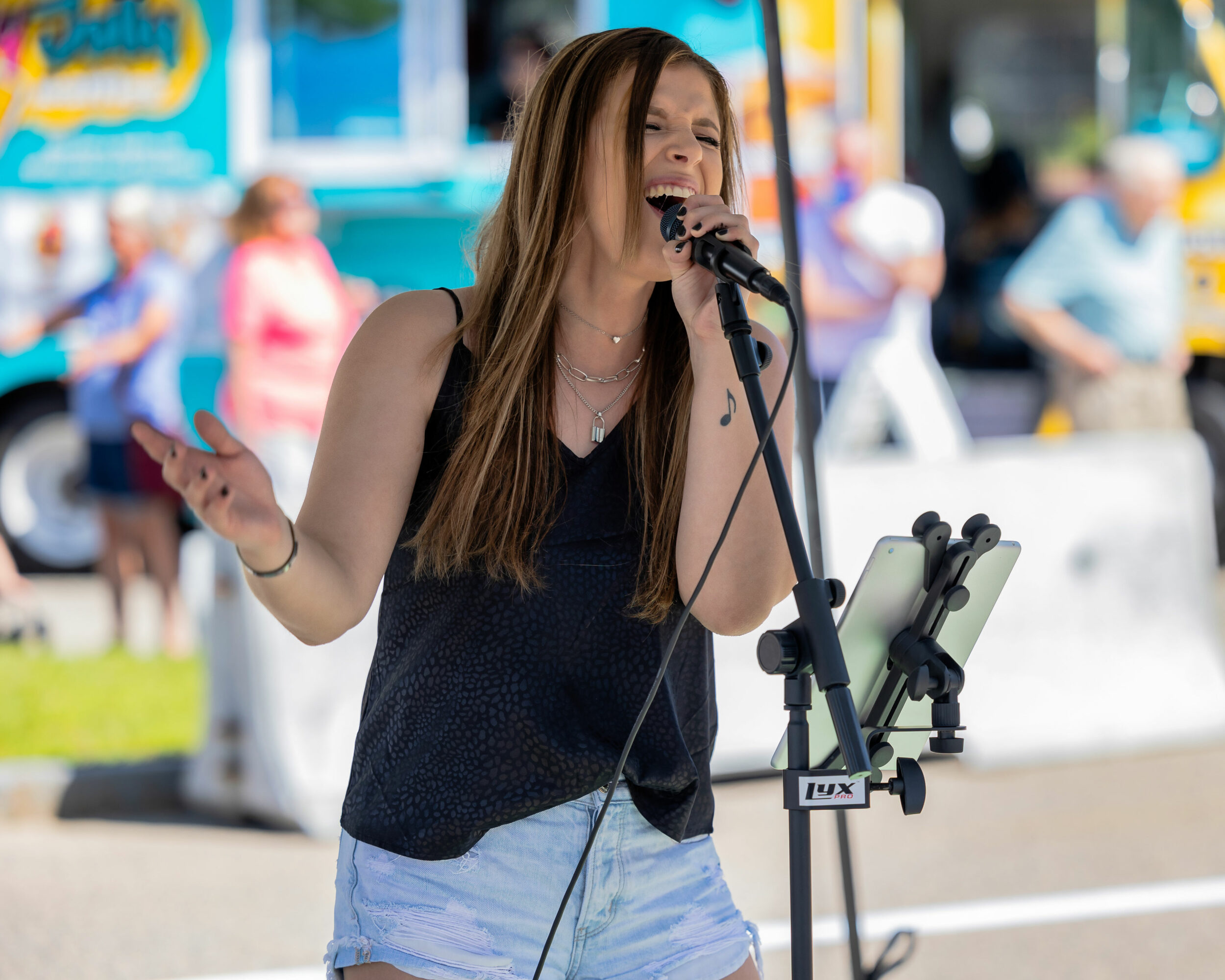 Young woman singing into microphone outdoors with people and colorful background.