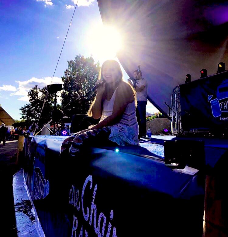 Person sitting on a stage with bright sunlight behind, trees, and stage equipment visible.