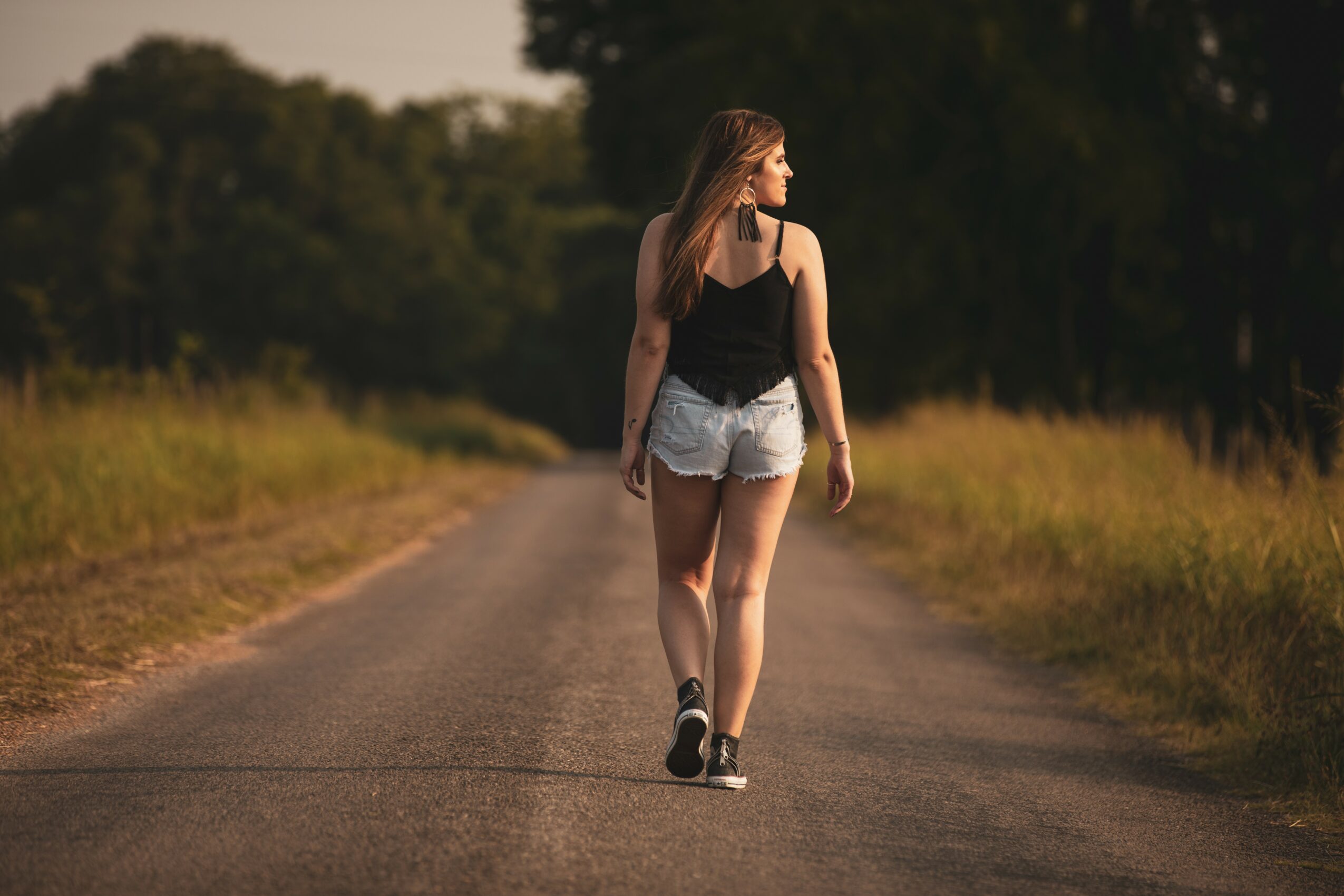 Young woman walking on a rural road with trees and grass on both sides, wearing a black top and denim shorts.