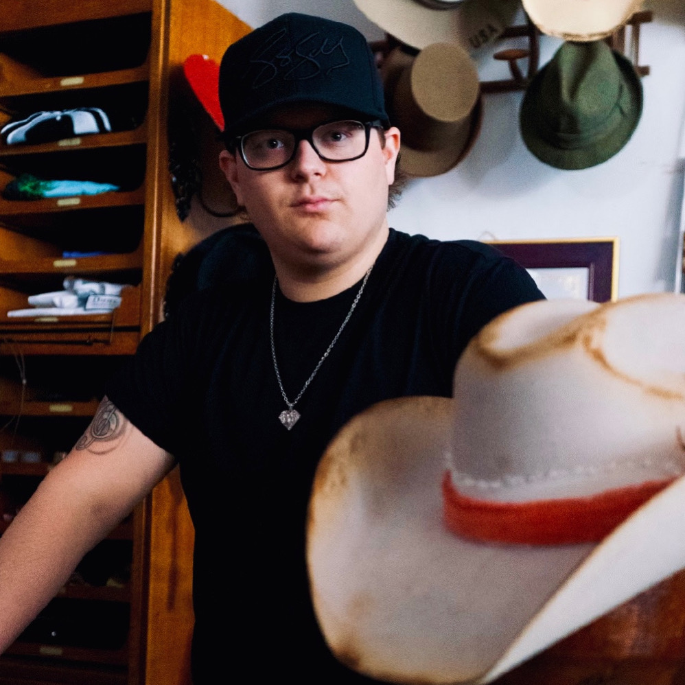 Person with glasses and a black cap, wearing a black shirt, standing indoors with hats on the wall behind.
