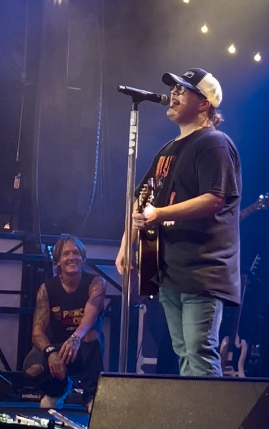 Person singing into a microphone on stage with another person sitting nearby, stage lights overhead.