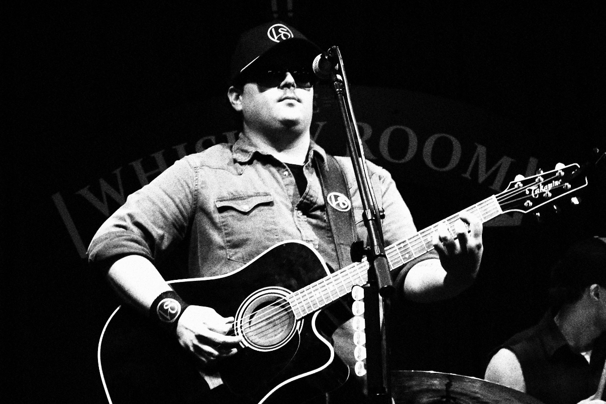 Person wearing sunglasses and a cap playing an acoustic guitar on stage with a microphone.