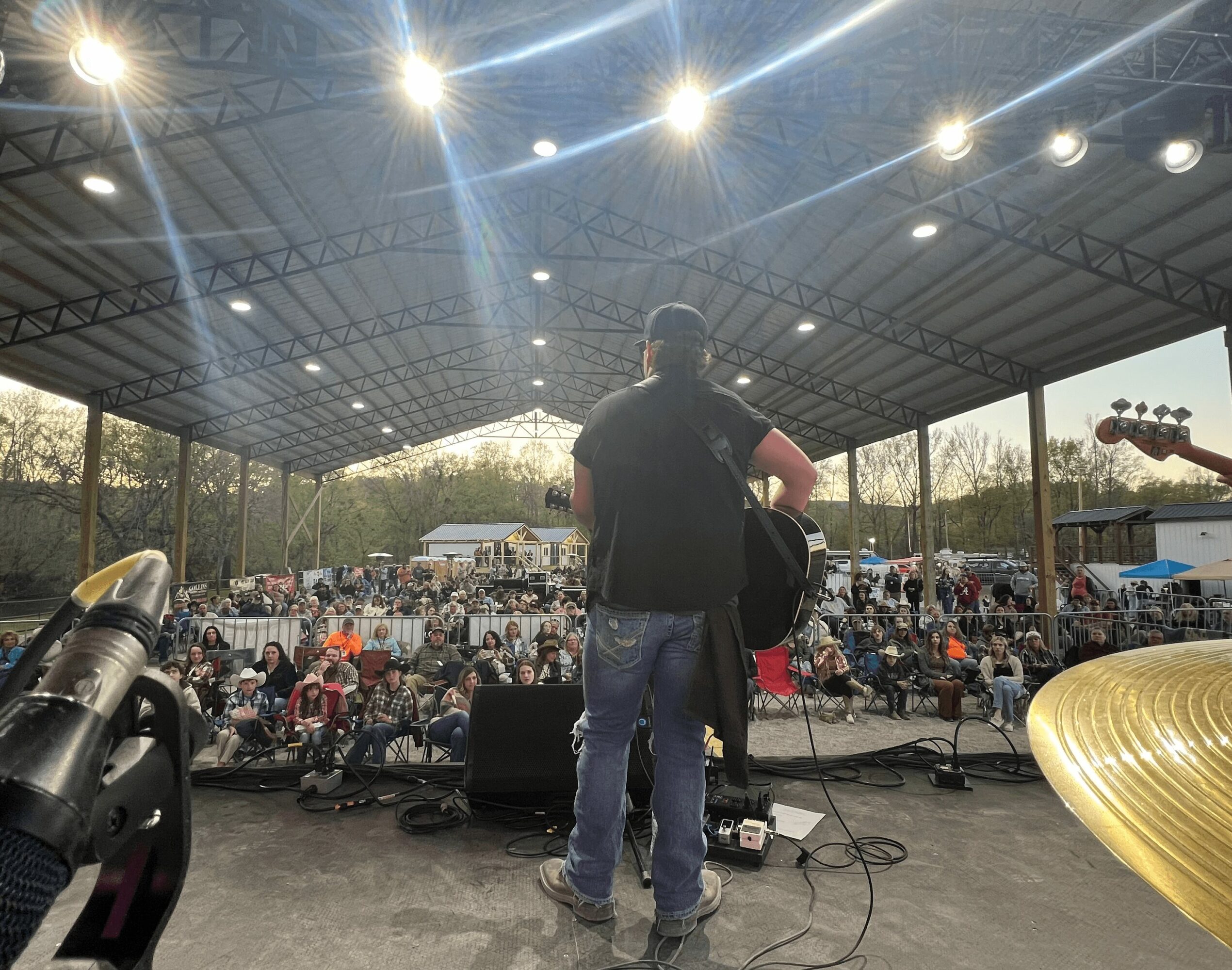Musician on stage with guitar performing in front of a large seated audience outdoors under a metal roof.