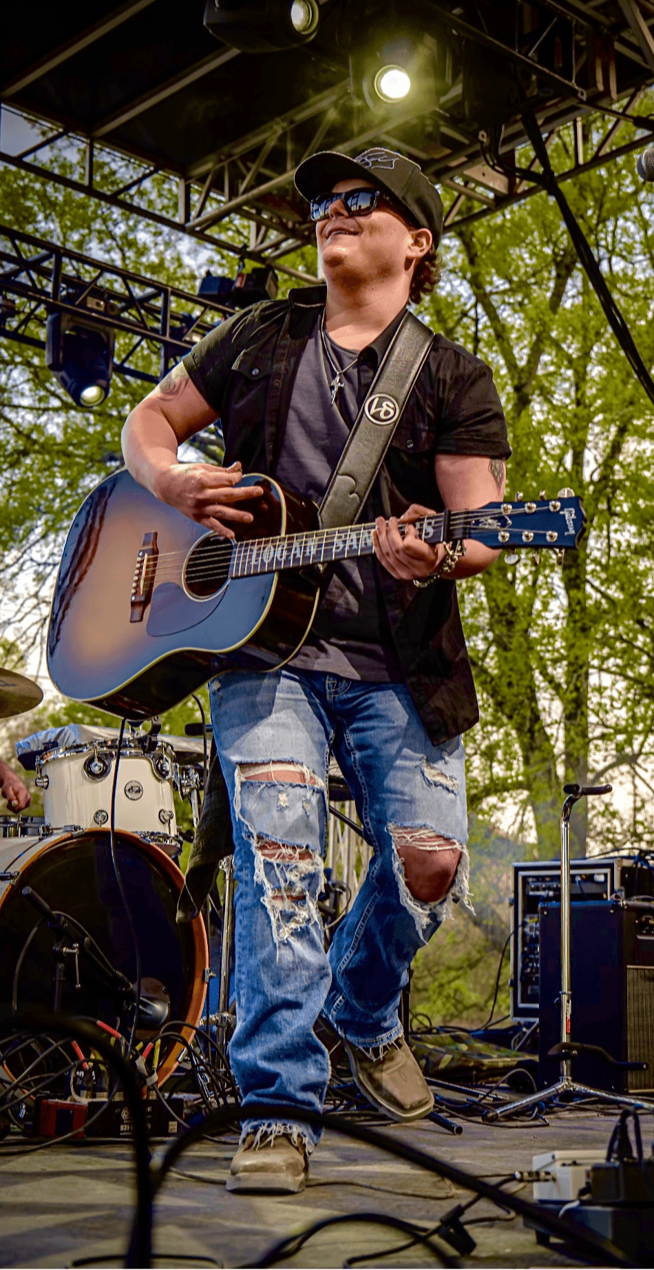 Person playing guitar on stage outdoors, wearing sunglasses, a hat, and ripped jeans, with musical equipment around.