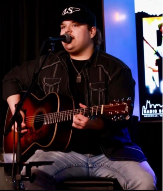 Person playing guitar and singing into microphone on stage, wearing black jacket and cap, with screen in background.