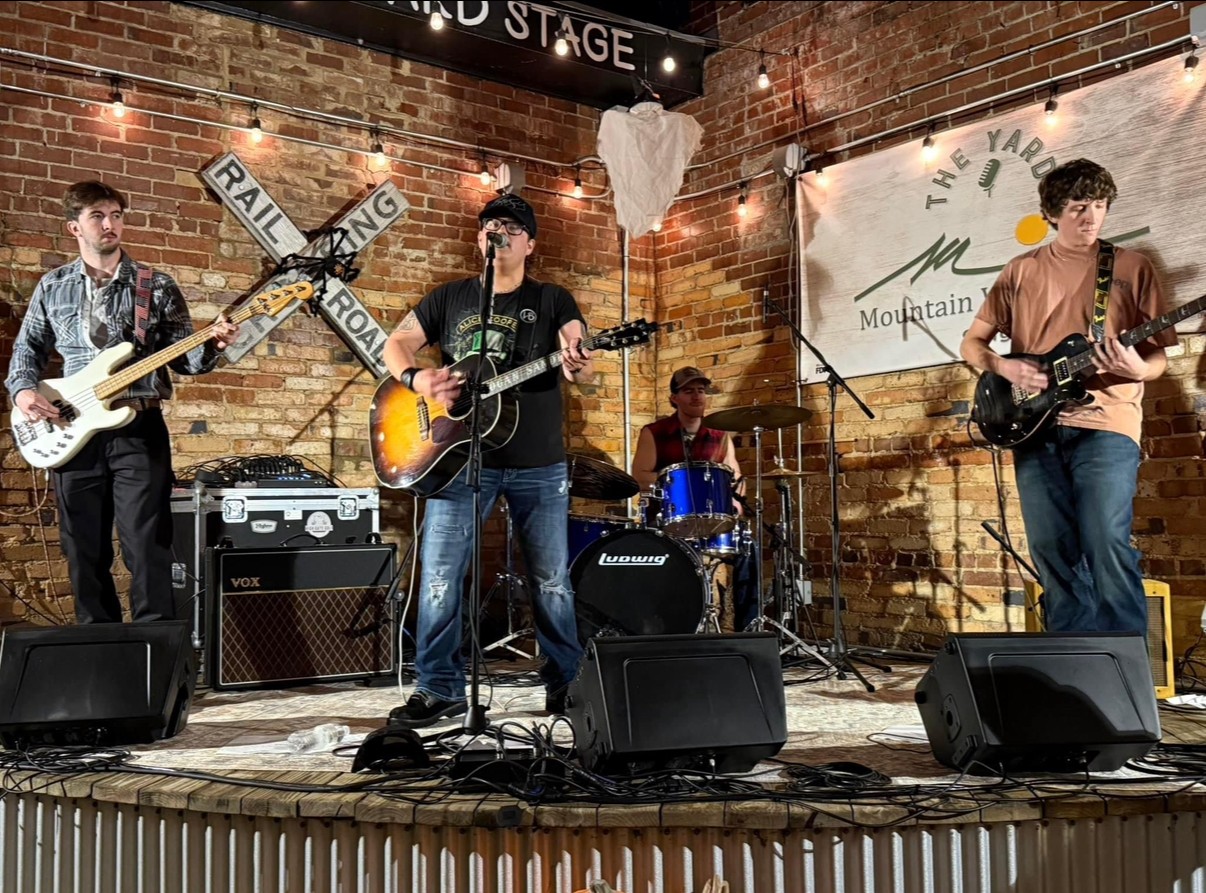 Four musicians perform on stage with guitars and drums, brick wall background, stage lights, and equipment visible.