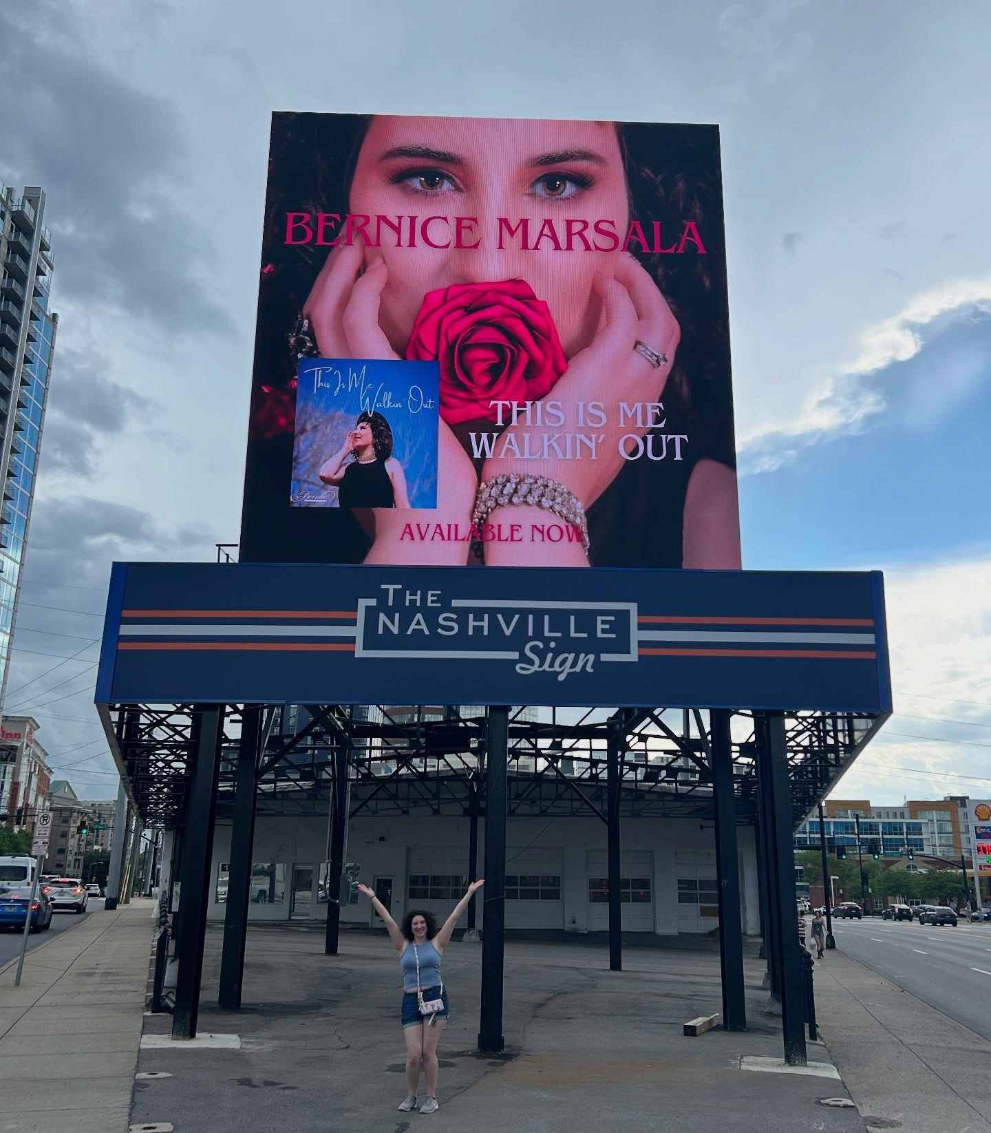Large billboard featuring a woman's face holding a pink rose, with text about Bernice Marsala and her album, against a city sky.