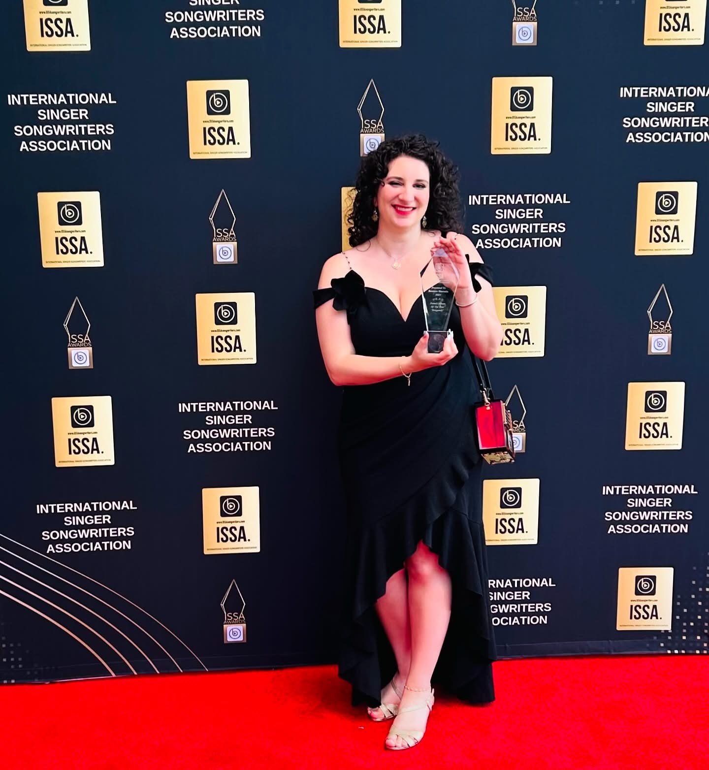 Woman in black dress holding an award on red carpet, backdrop with logos behind her.