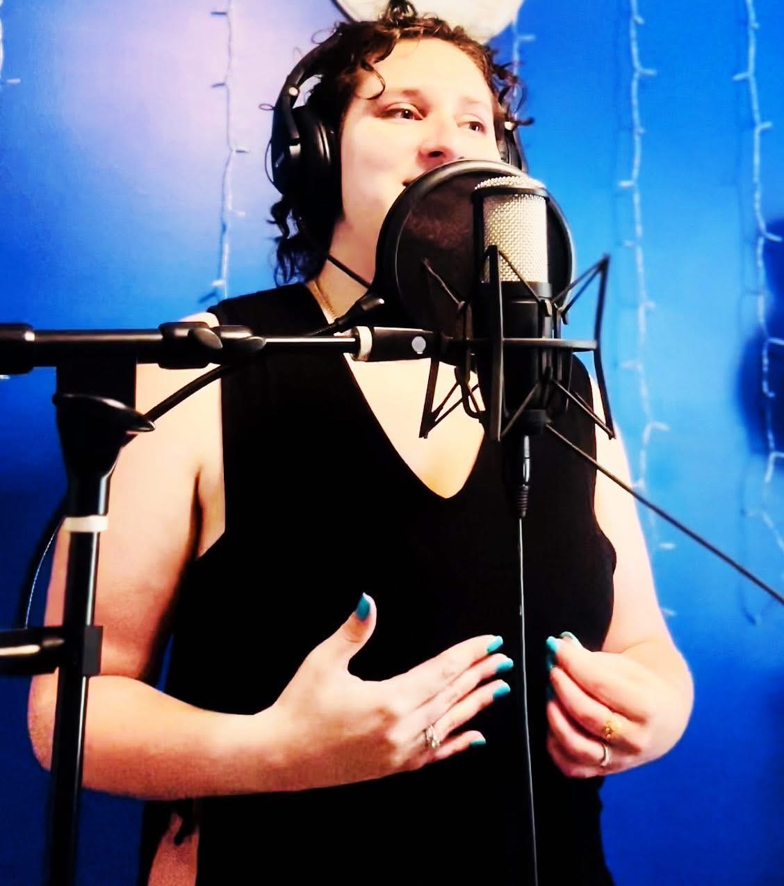 Woman singing into a microphone with headphones in a recording studio, blue background, wearing a black sleeveless top.