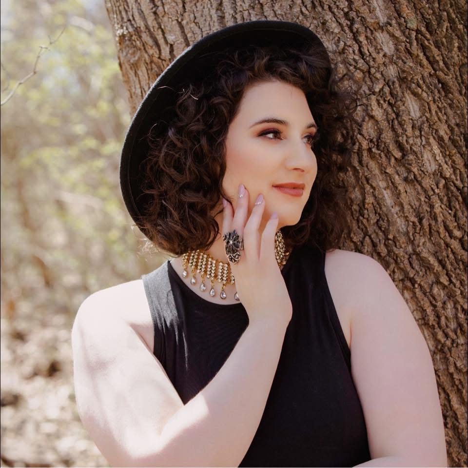 Woman with curly hair wearing a black hat, sleeveless black top, and jewelry, leaning against a tree outdoors.