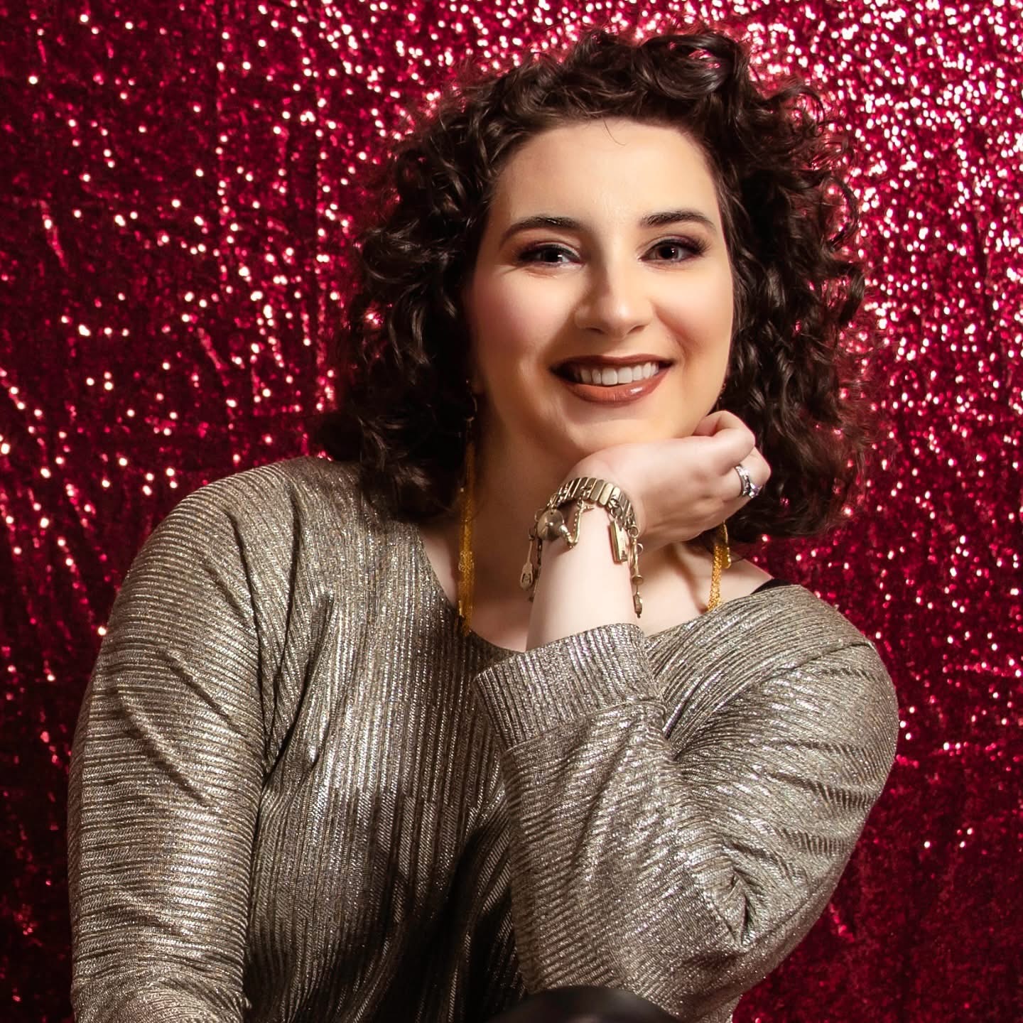 Young woman with curly hair smiling, resting her chin on her hand, against a red glittery background.