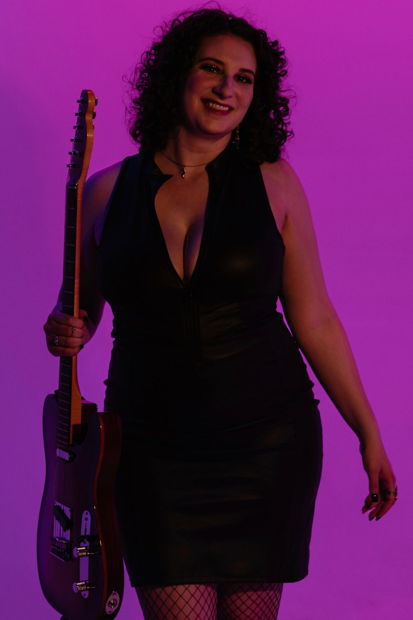 Woman with curly hair holding a guitar, smiling, wearing a black dress, against a purple background.