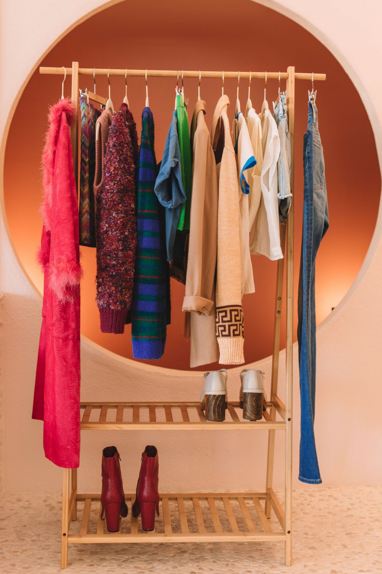 Clothing rack with colorful dresses, shirts, and pants, and a pair of boots on the bottom shelf, inside a circular frame.