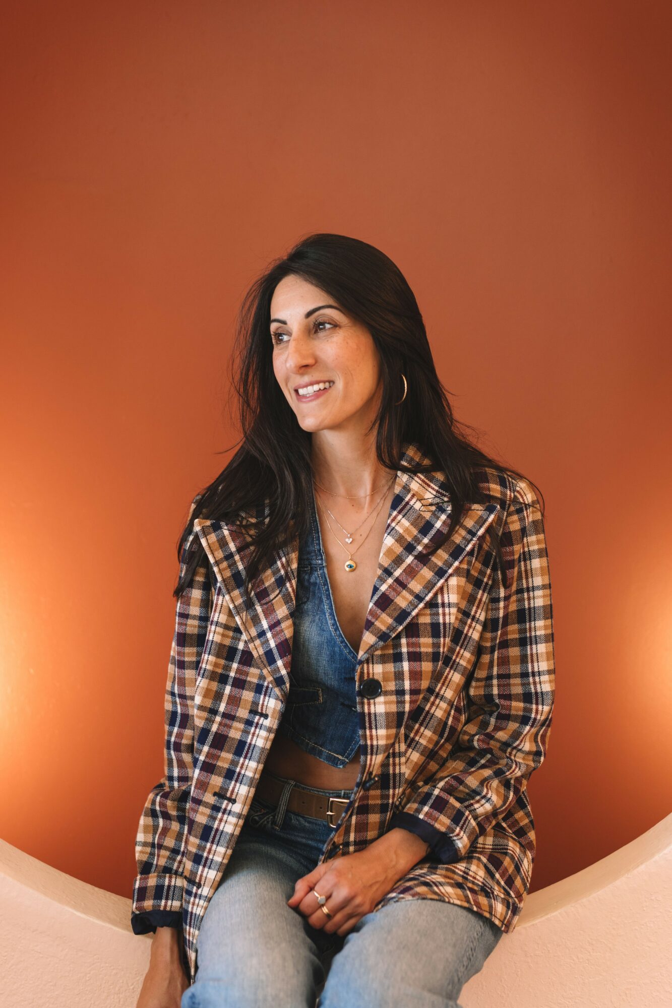 Woman with long dark hair smiling, wearing a plaid blazer and jeans, sitting against an orange background.