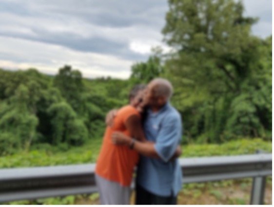 Two people hugging outdoors near a guardrail with trees and cloudy sky in background.