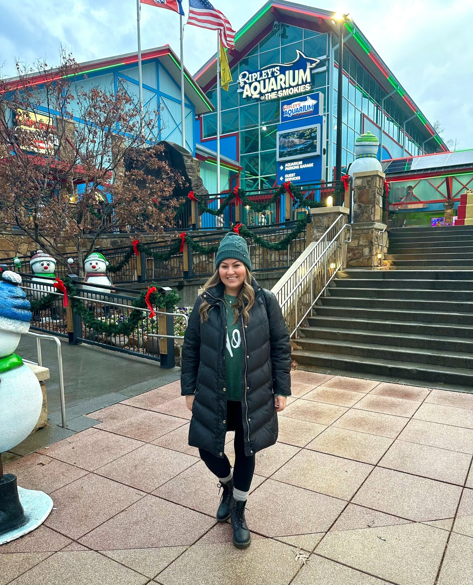 Young woman standing outside Oceanarium entrance, smiling, wearing a beanie, coat, and boots, with stairs and colorful building behind.