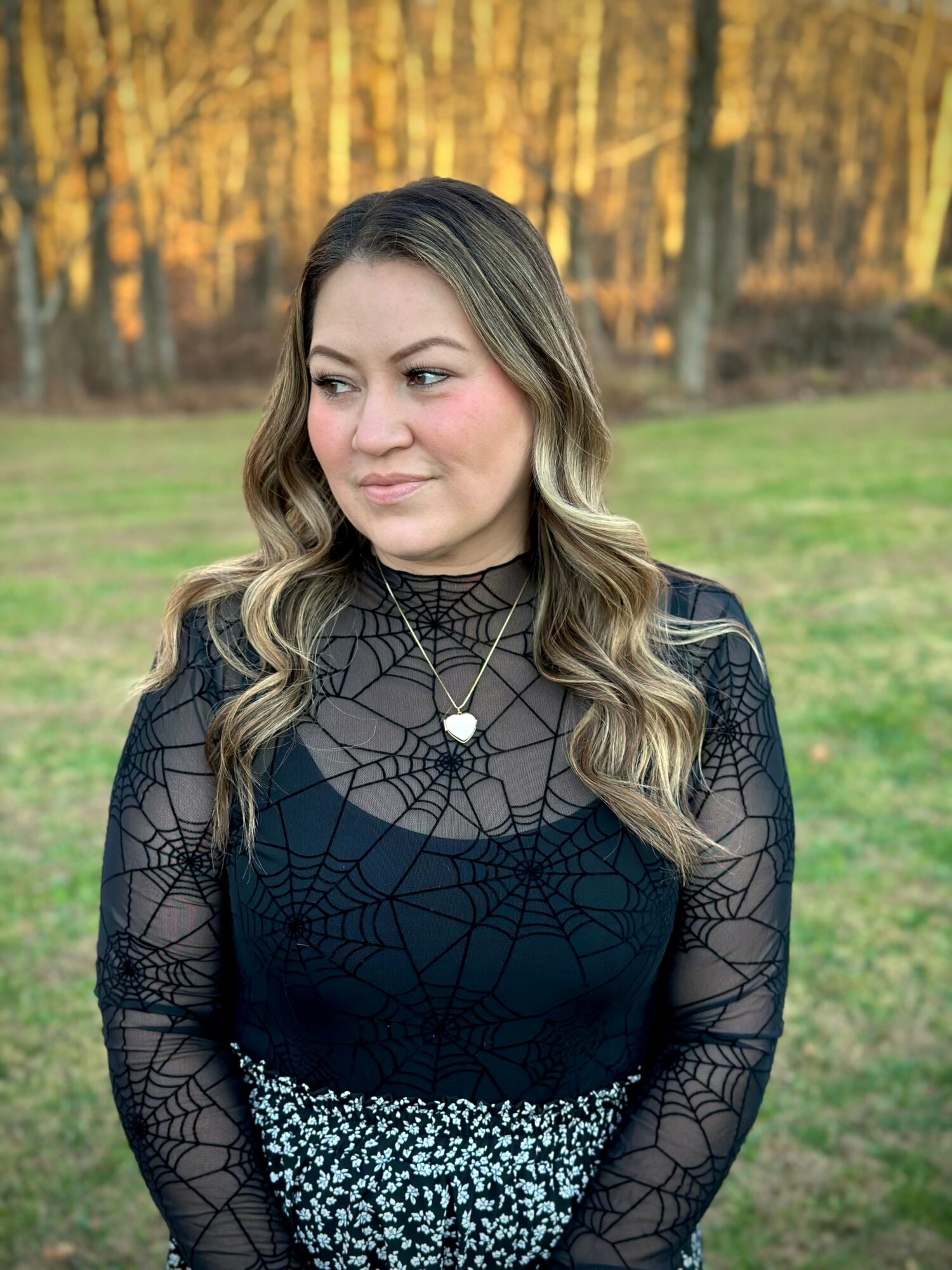 Young woman with wavy hair standing outdoors in a park with trees and grass, wearing a black lace top and layered necklaces.