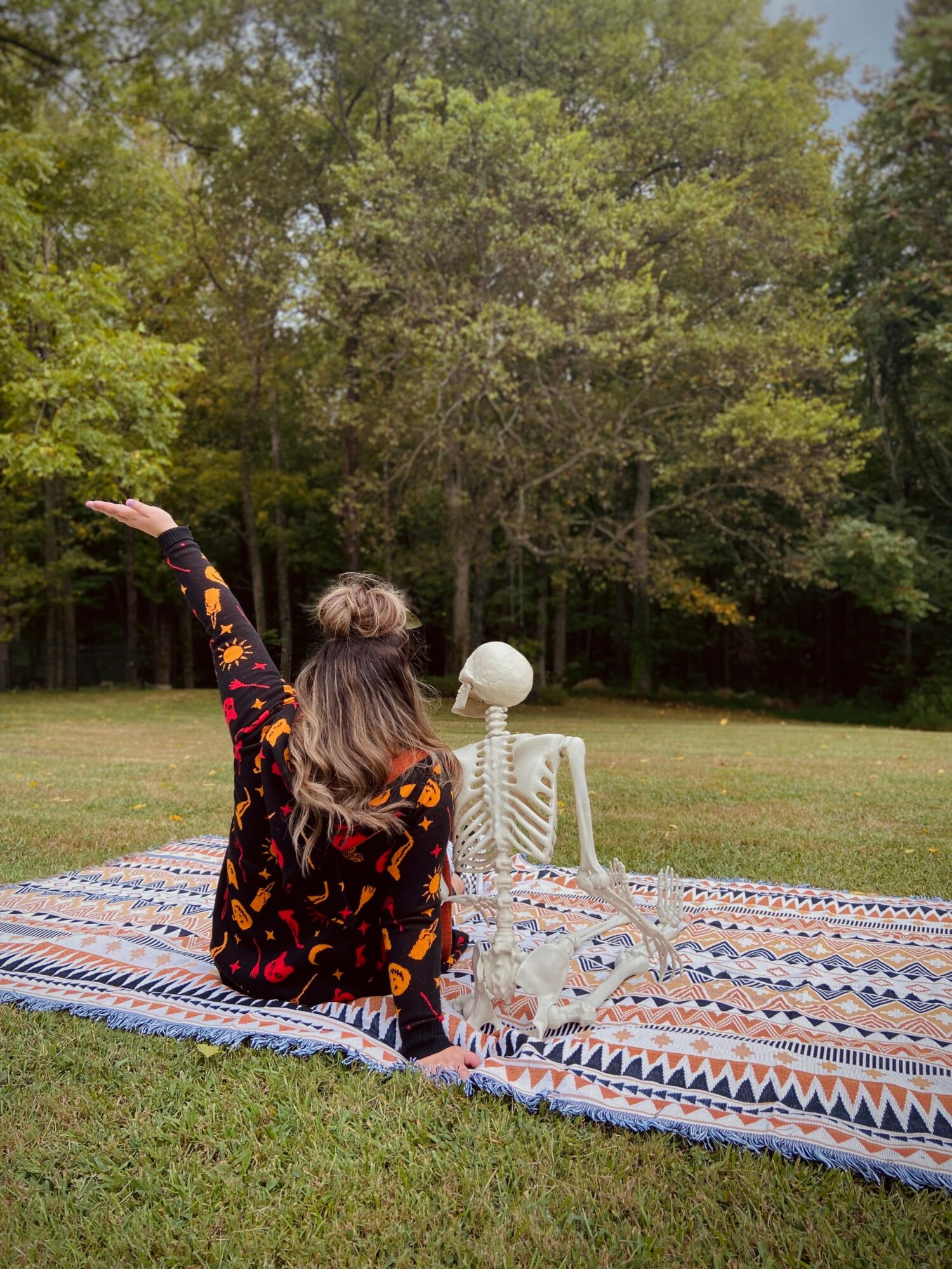 Child sitting on a patterned blanket outdoors, reaching toward a skeleton model with trees in the background.