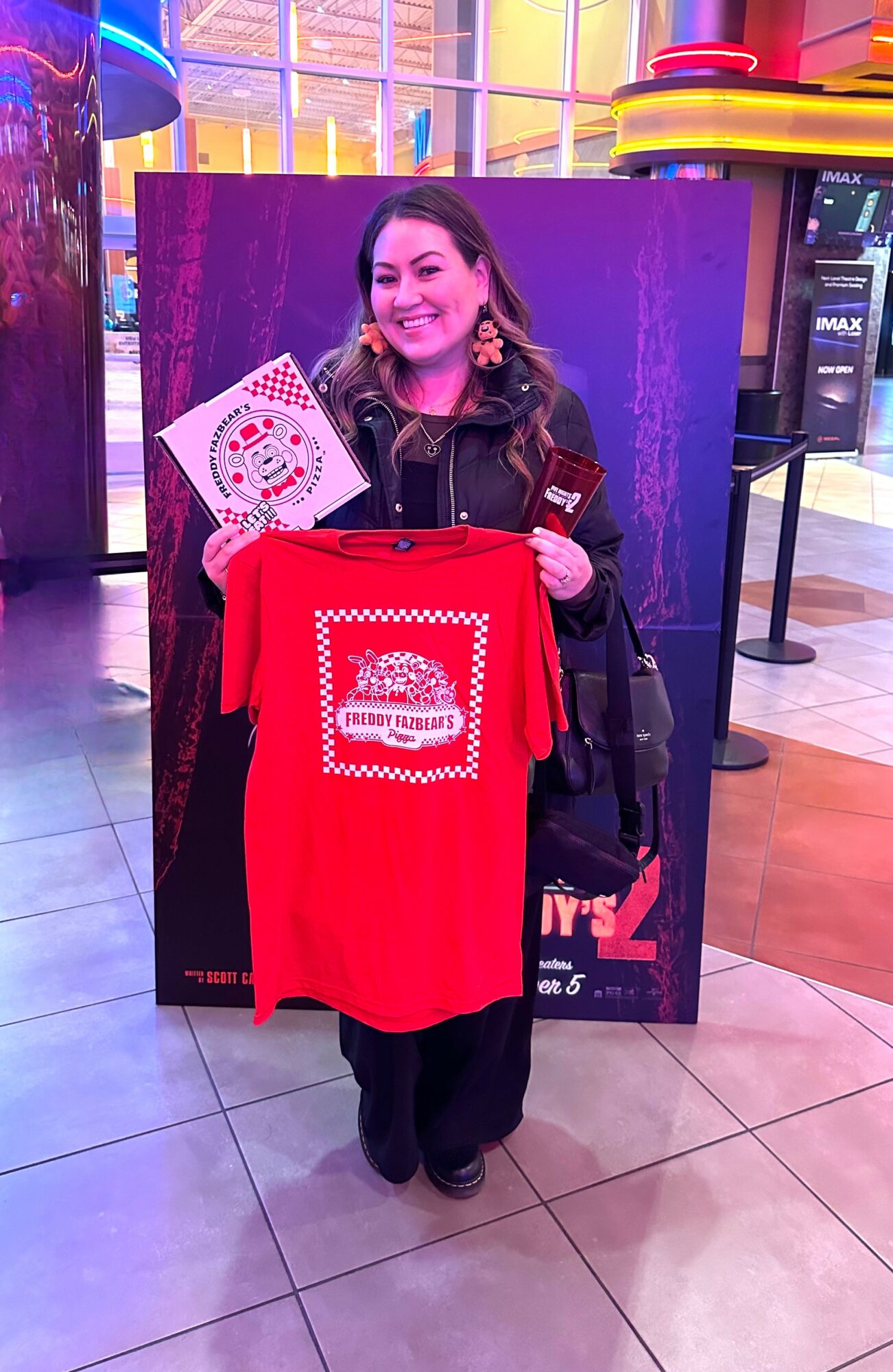 Woman standing indoors, holding a red T-shirt with a design, smiling, with colorful background and shopping bags.