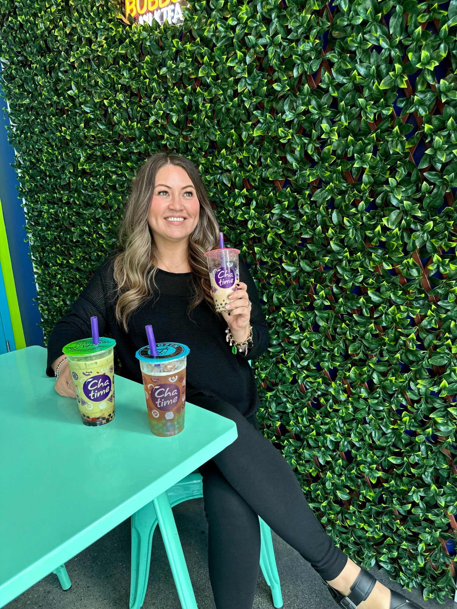 Woman sitting at a table with colorful drinks, smiling, against a green leafy wall background.