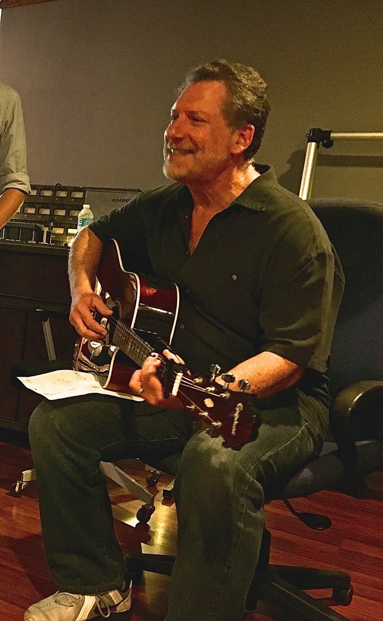 Man sitting on a chair playing an acoustic guitar, smiling, in a room with music equipment.