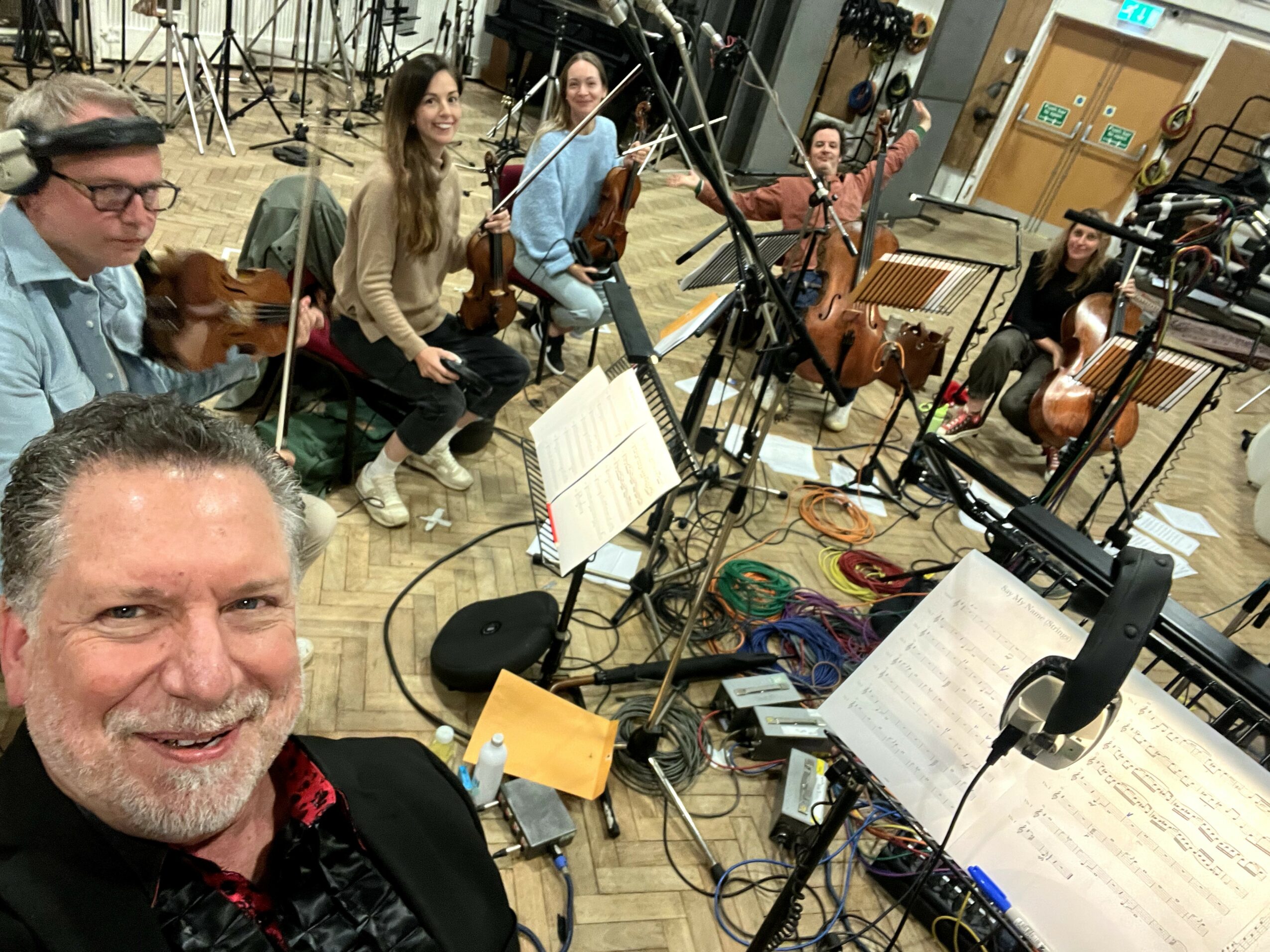 Group of musicians in a recording studio, with a man in the foreground smiling at the camera, music stands, and instruments.