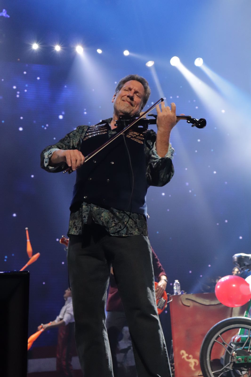 Musician playing violin on stage with blue and white lights, wearing dark clothing and glasses.