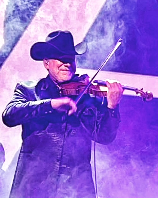 Person wearing a large hat and leather jacket playing a violin on stage with purple lighting.